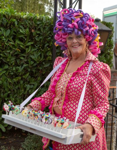 De Candy Lady van Uw Chef Catering in kleurrijk kostuum met krul-lintenpruik draagt een tray met zoete lekkernijen op Piet's 70 party. Foto: AbDonkerFotografie.